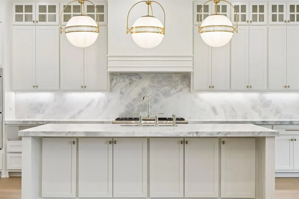 Bright, all-white kitchen with marble backsplash and island. Three globe pendant lights with gold accents hang above the island.