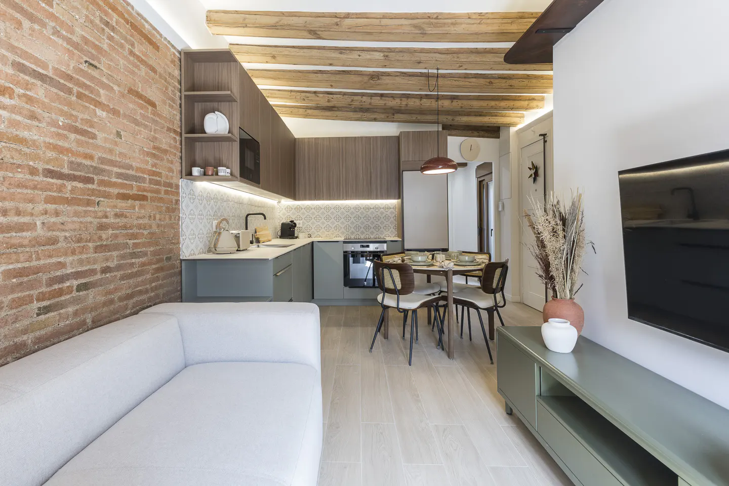 Open-concept living space with exposed brick wall, white sofa, kitchen with wood cabinets, and dining table with four chairs.