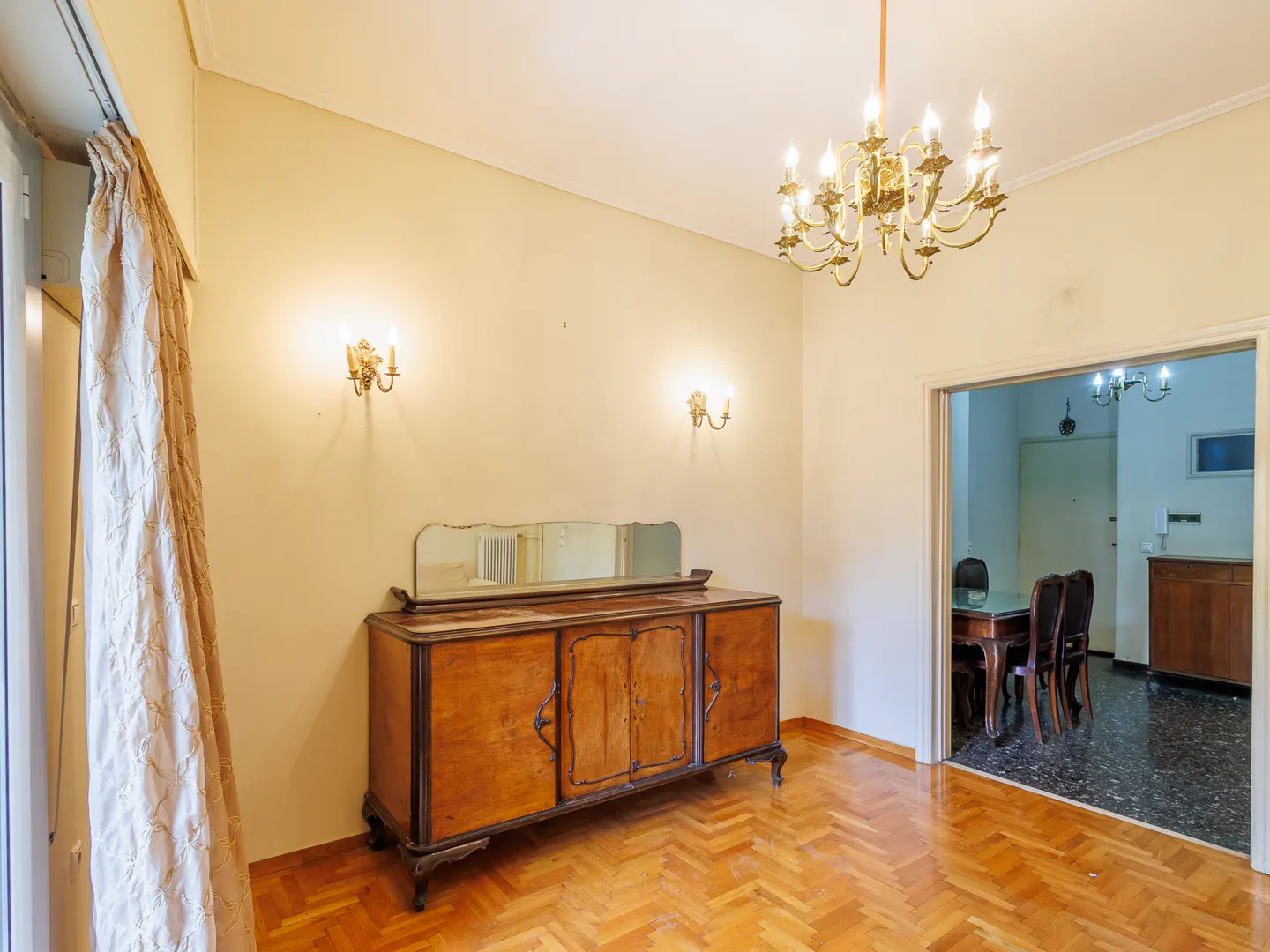 A room with parquet floors, a wooden sideboard with a mirror, and a gold chandelier. A dining room is visible through the doorway.