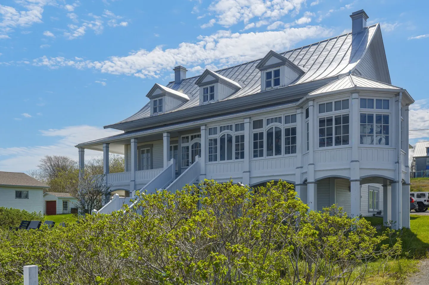 A large, white two-story coastal home with a metal roof and wrap-around porch, partially obscured by green bushes.