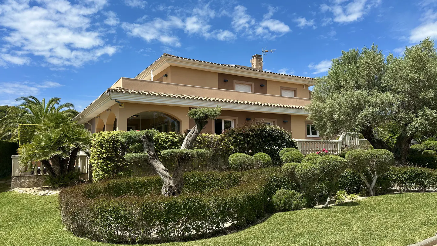 Two-story tan house with a tile roof, green lawn, and manicured hedges under a blue sky.