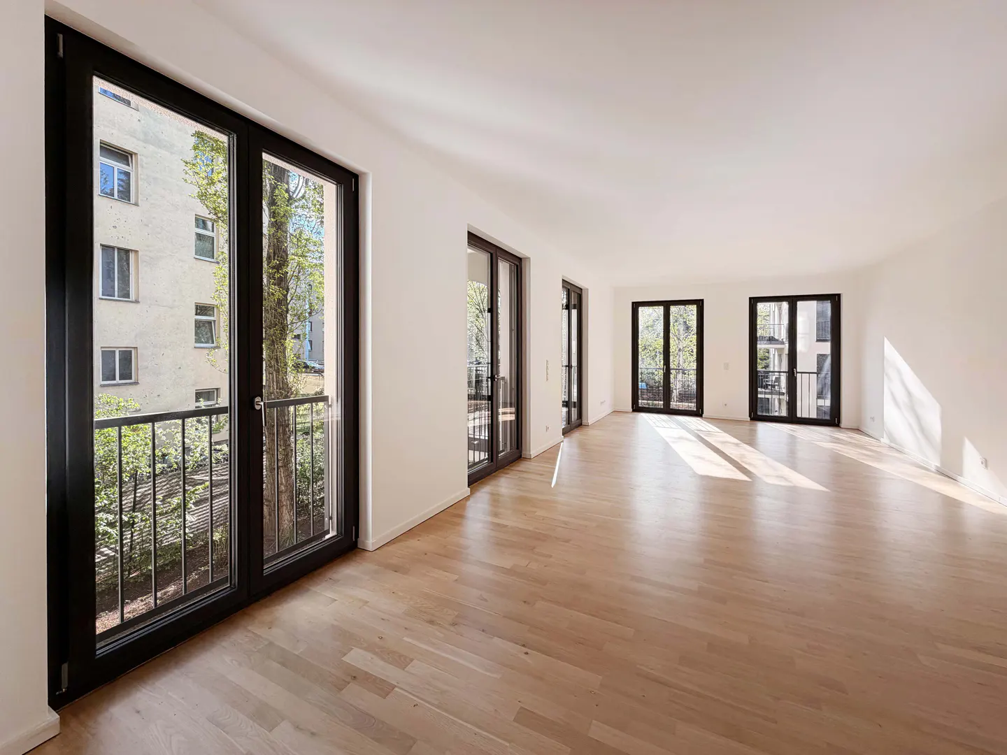 Bright, empty room with light wood floors and white walls. Black-framed glass doors lead to balconies. Sunlight streams in.