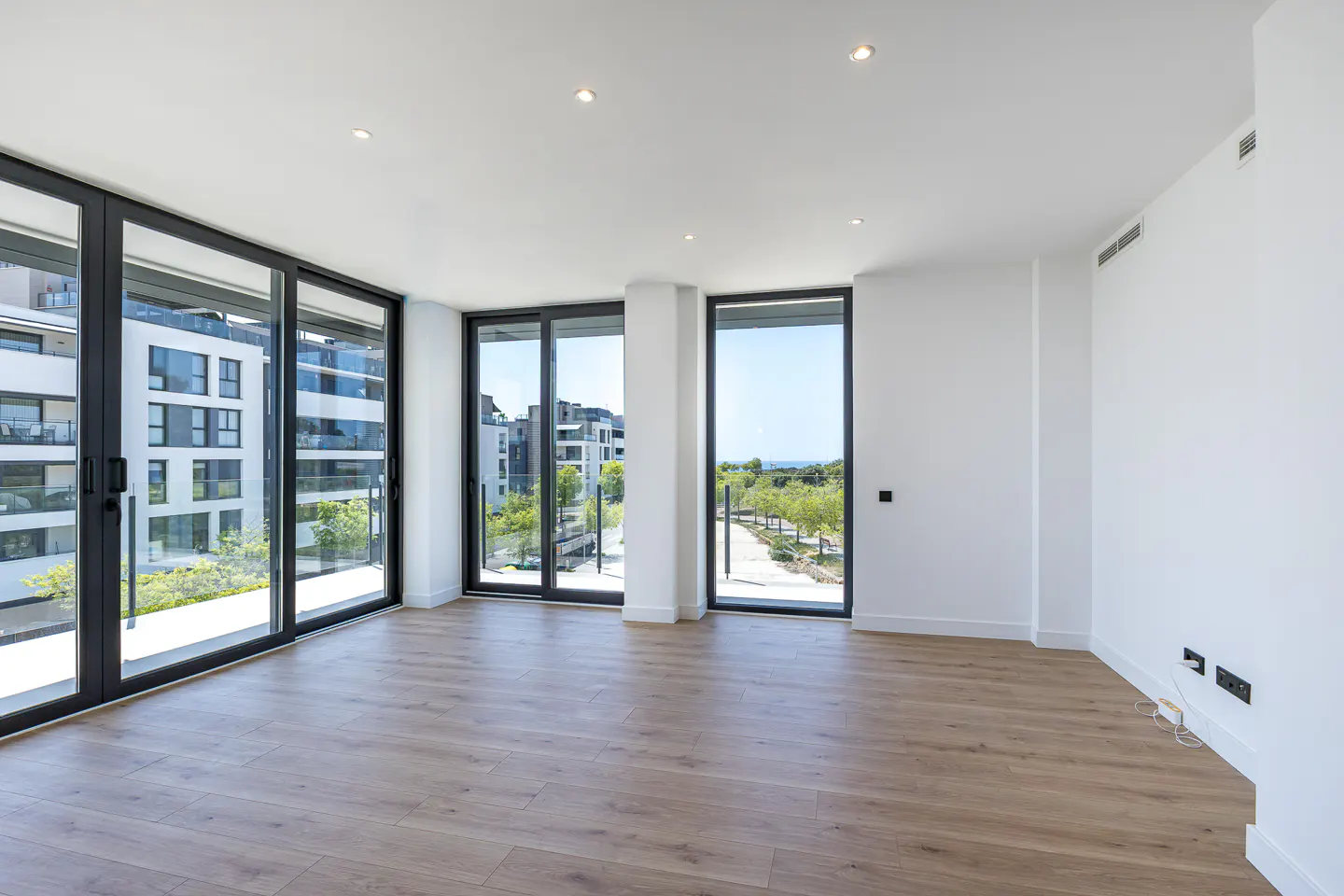 Bright, empty room with light wood floors, white walls, and black-framed sliding glass doors to a balcony with city views.