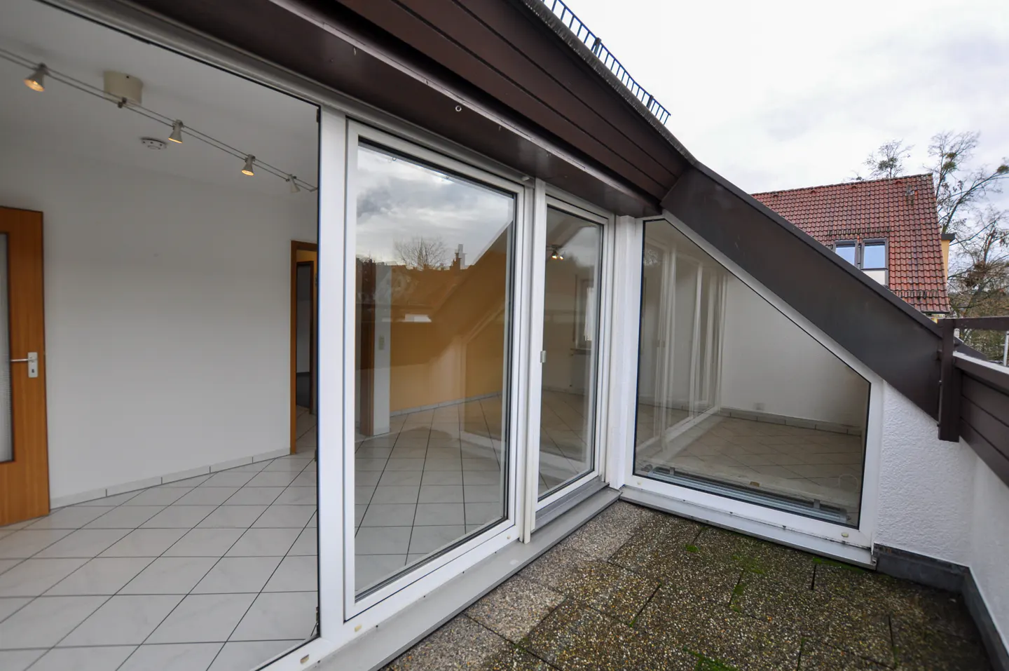 A bright room with white tile floors opens to a balcony through large glass doors. The balcony has stone tiles.
