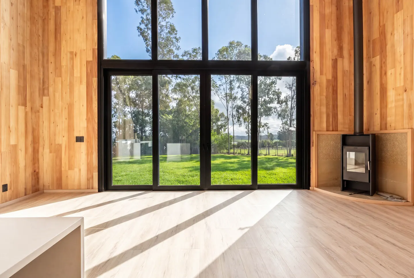 Modern room with light wood walls, floor-to-ceiling windows, a black fireplace, and a view of a sunny green backyard.
