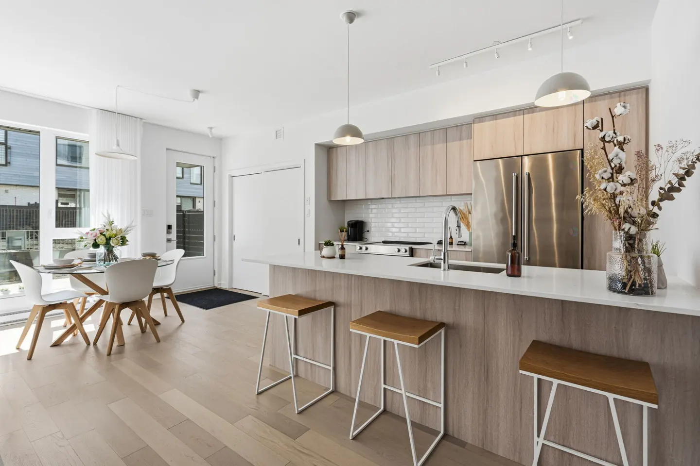 Bright, modern kitchen and dining area with wood floors, white walls, and stainless steel appliances. A glass table with white chairs sits near a window.