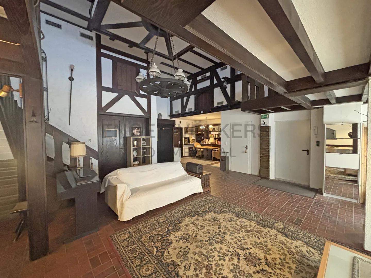 Interior view of a home with exposed beams, a white sofa, and a large rug on a brick floor. A staircase and a sword on the wall are visible.
