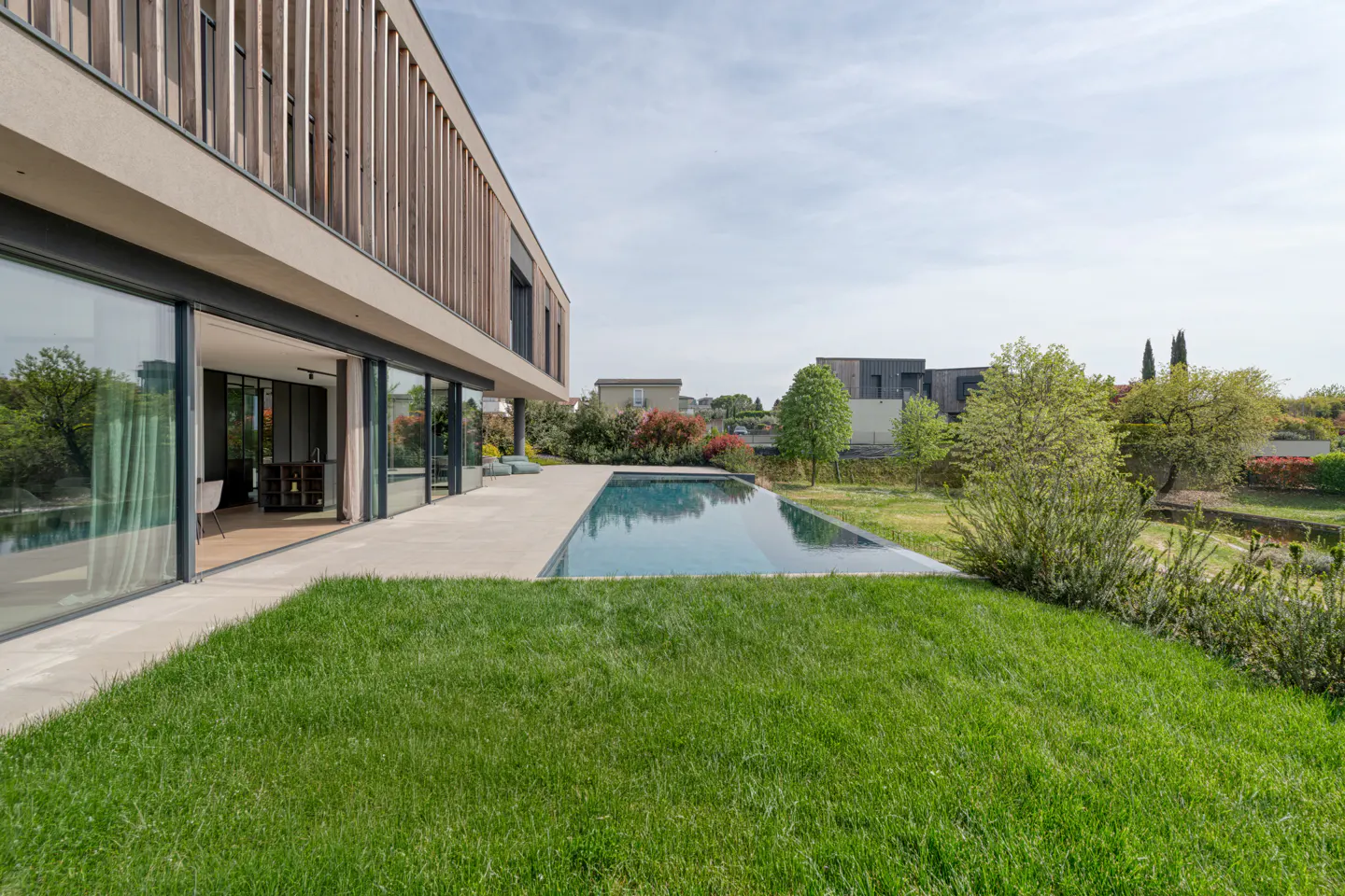 Modern home with a pool. The house has large glass doors and a wood slat facade. Green grass in the foreground.
