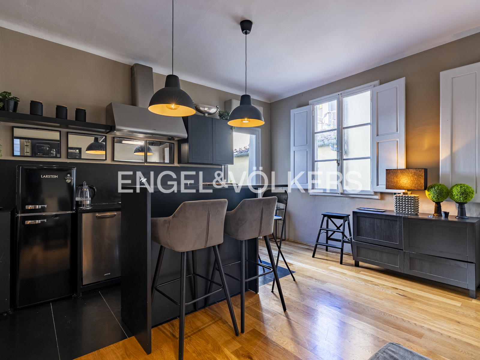 A modern kitchen with black cabinets, wood floors, and two pendant lights. Two stools are at the counter. A window with white shutters is in the background. [AI-generated text]