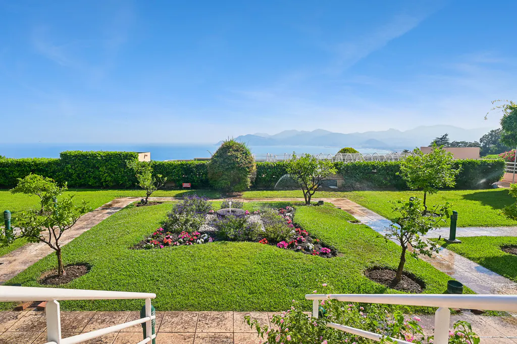 Lush garden with a central flower bed and fountain, overlooking a scenic ocean view with distant mountains under blue sky.
