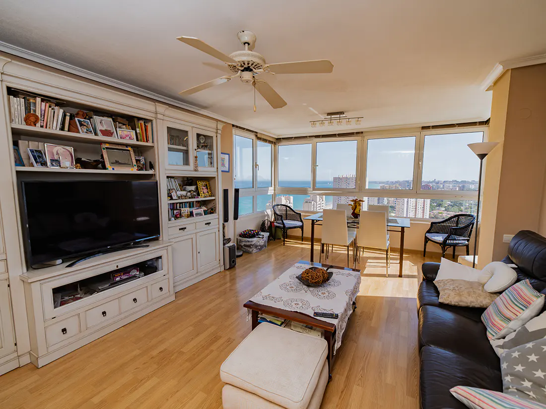 Bright living room with wood floors, a white built-in bookcase, a black leather sofa, and a dining area with a view of the ocean.