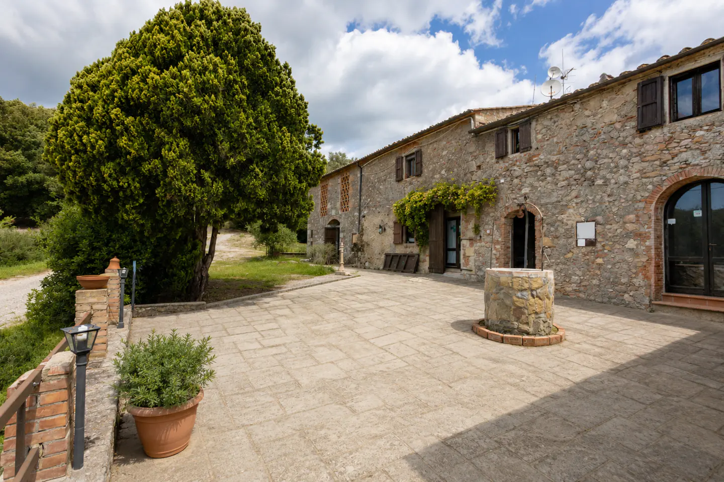 Exterior view of a stone house with brown shutters, a stone well, and a large green tree.