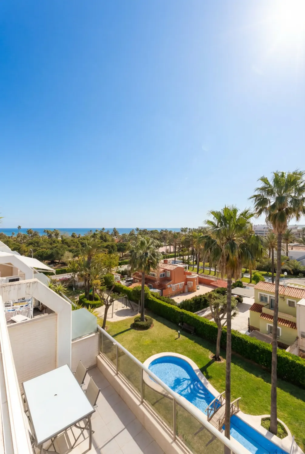 Balcony view of a resort with a pool, palm trees, and the ocean under a clear blue sky. Table and chairs on the balcony.