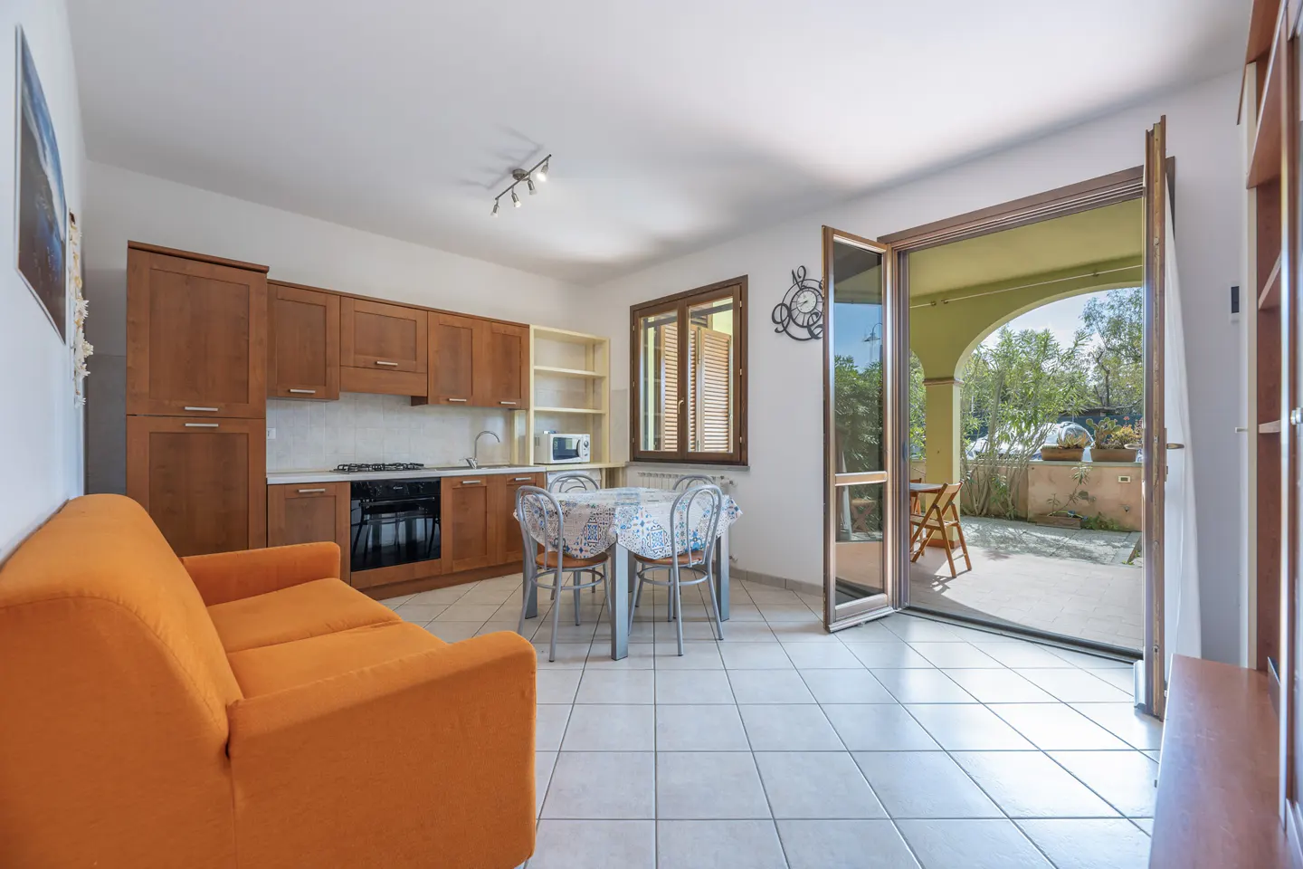 Bright apartment interior with orange sofa, wood cabinets, dining table, and open doors to a patio.