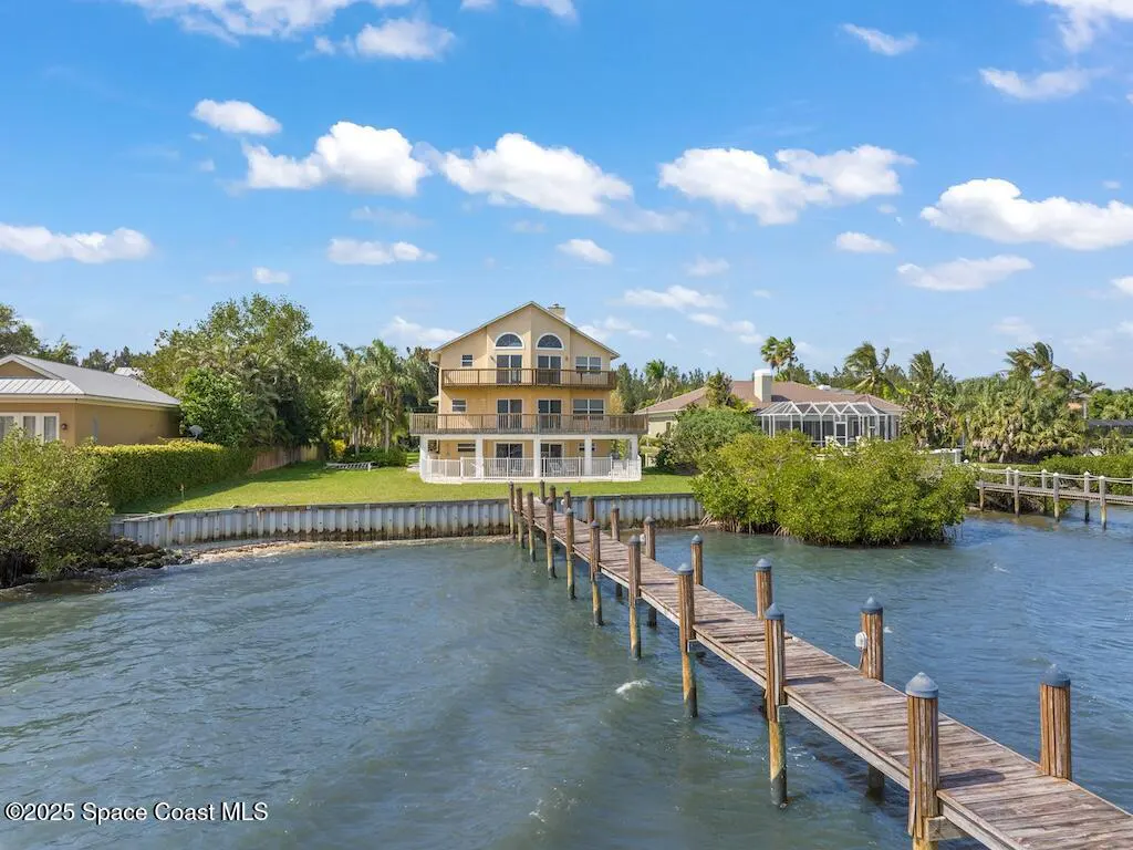 Three-story yellow waterfront home with a long wooden dock extending into the water under a bright blue sky.
