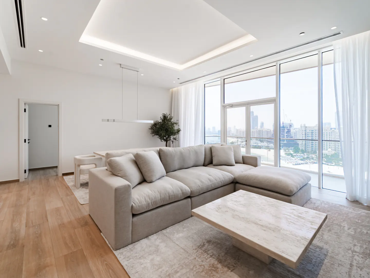 Bright, modern living room with a beige sectional sofa, marble coffee table, and floor-to-ceiling windows with a city view.