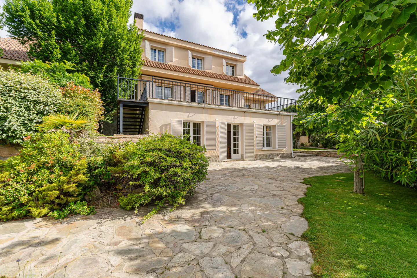 Exterior view of a three-story beige house with white shutters, a black iron balcony, and a stone patio surrounded by lush greenery.