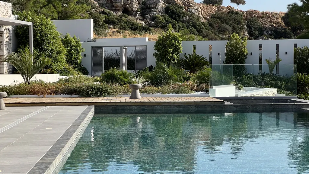 A modern pool with clear water reflects the sky. A wooden deck and lush greenery surround the pool, with a white building and hillside in the background.