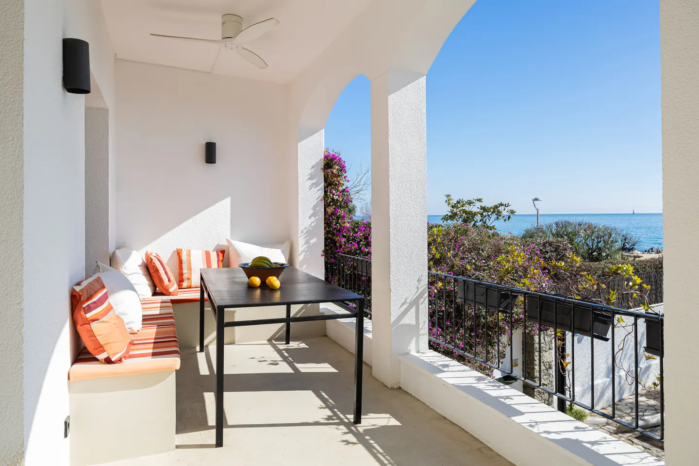Covered patio with built-in bench, striped cushions, black dining table, and ocean view framed by white arches.