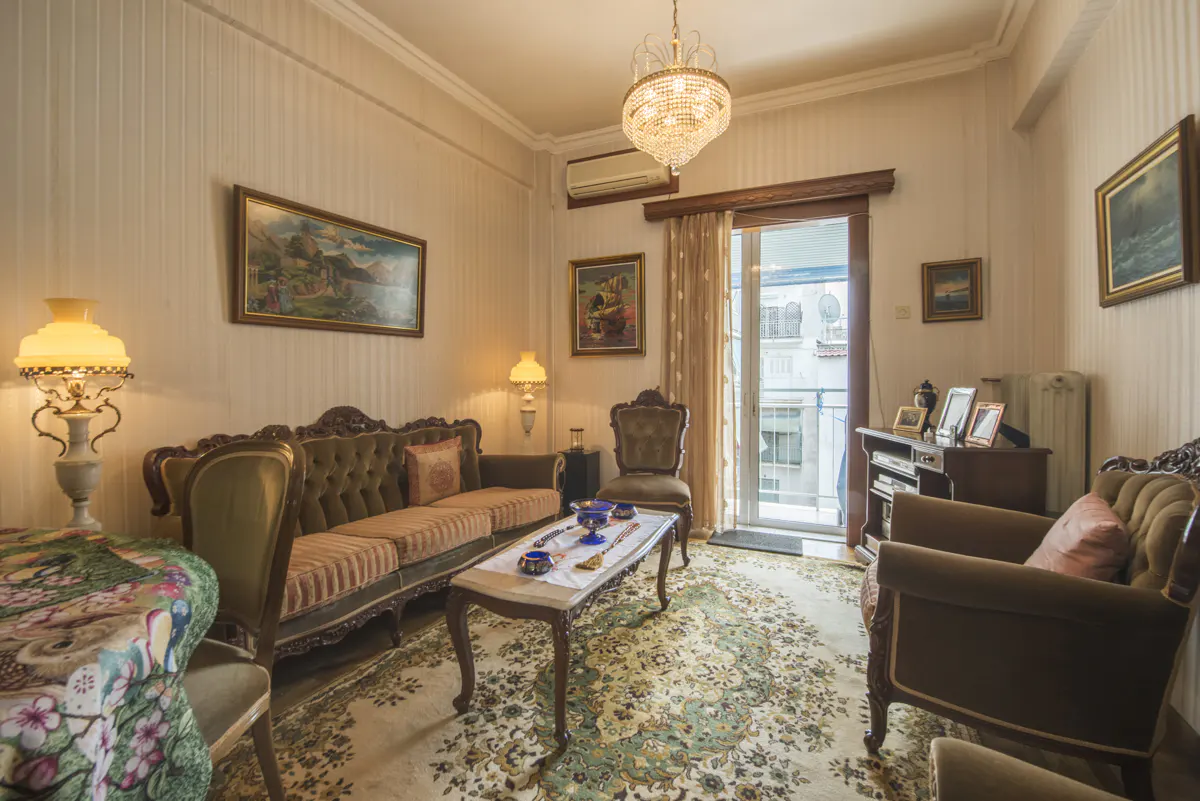 Ornate living room with olive sofa, floral rug, and crystal chandelier. Balcony door open to city view.