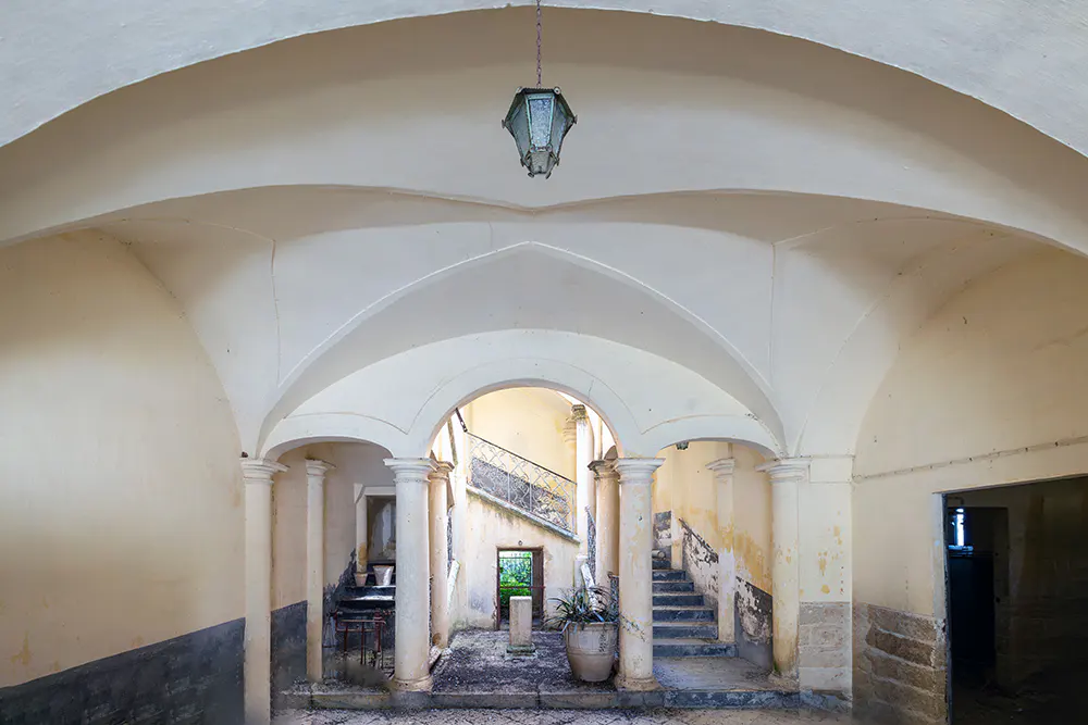 Arched hallway with columns leads to a staircase. A hanging lantern illuminates the cream-colored walls and stone floor.