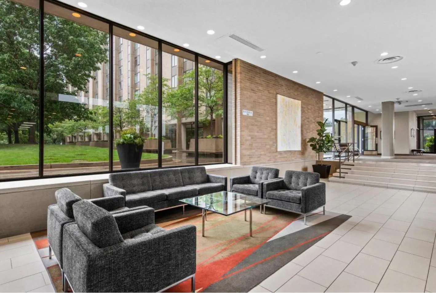 Lobby with gray sofas and chairs around a glass table on a colorful rug. Large windows overlook a green lawn and building.