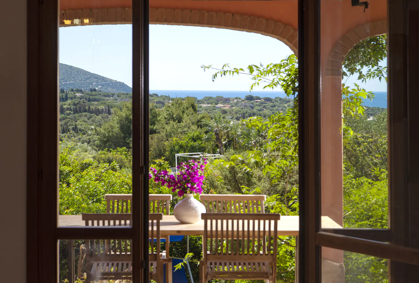 View through arched doorway to patio with table, chairs, and flowers. Lush green trees and blue ocean in background.