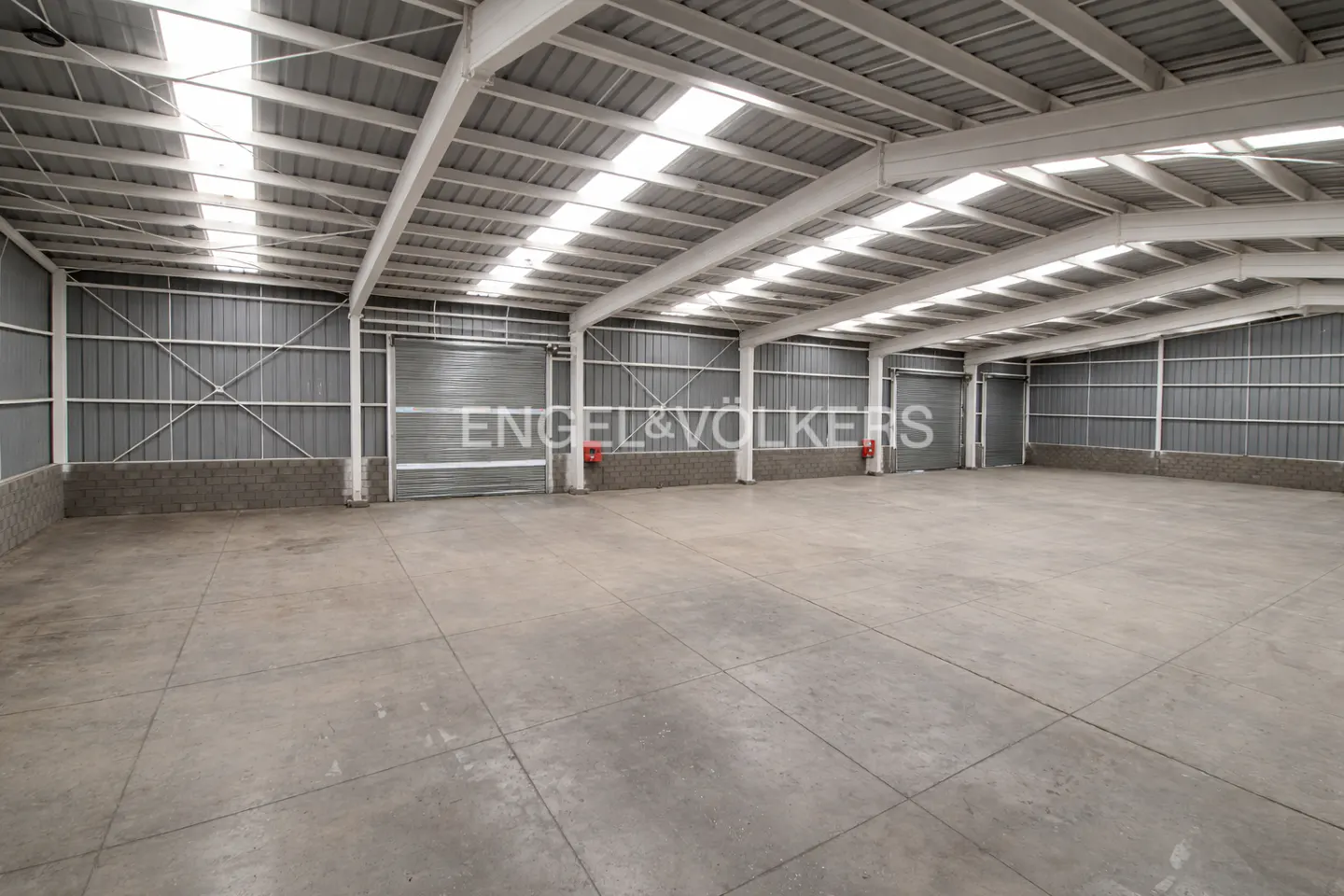 Large, empty industrial warehouse with concrete floors, metal walls, and skylights in the high ceiling.