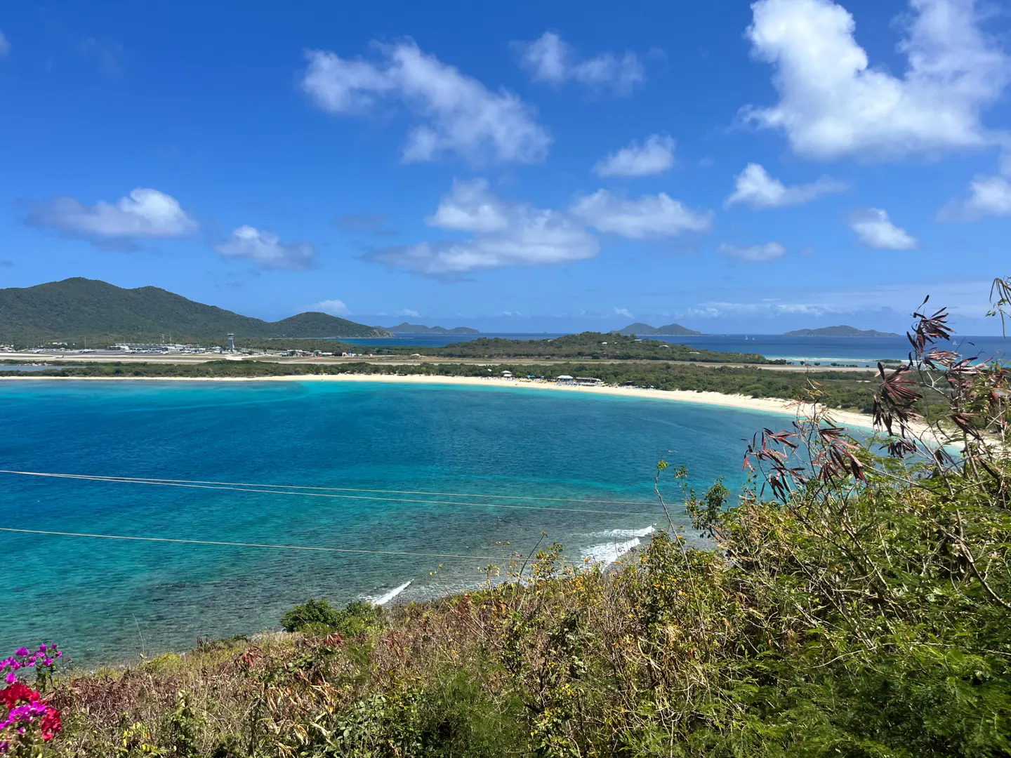 Scenic view of a turquoise bay with a white sand beach, green hills, and a blue sky with scattered clouds. Lush vegetation in the foreground.