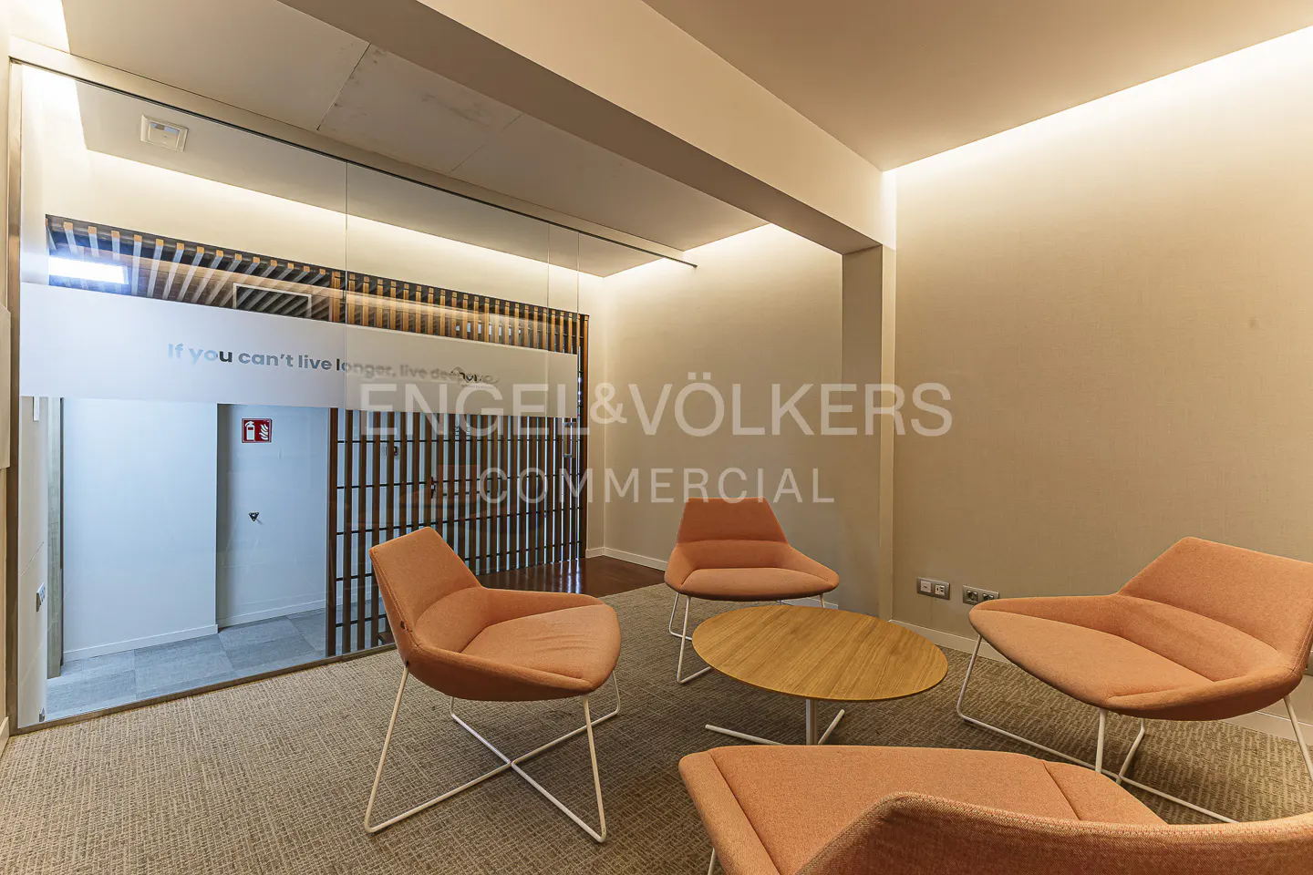 A modern office waiting area with orange chairs, a wood table, and a glass-enclosed entrance.