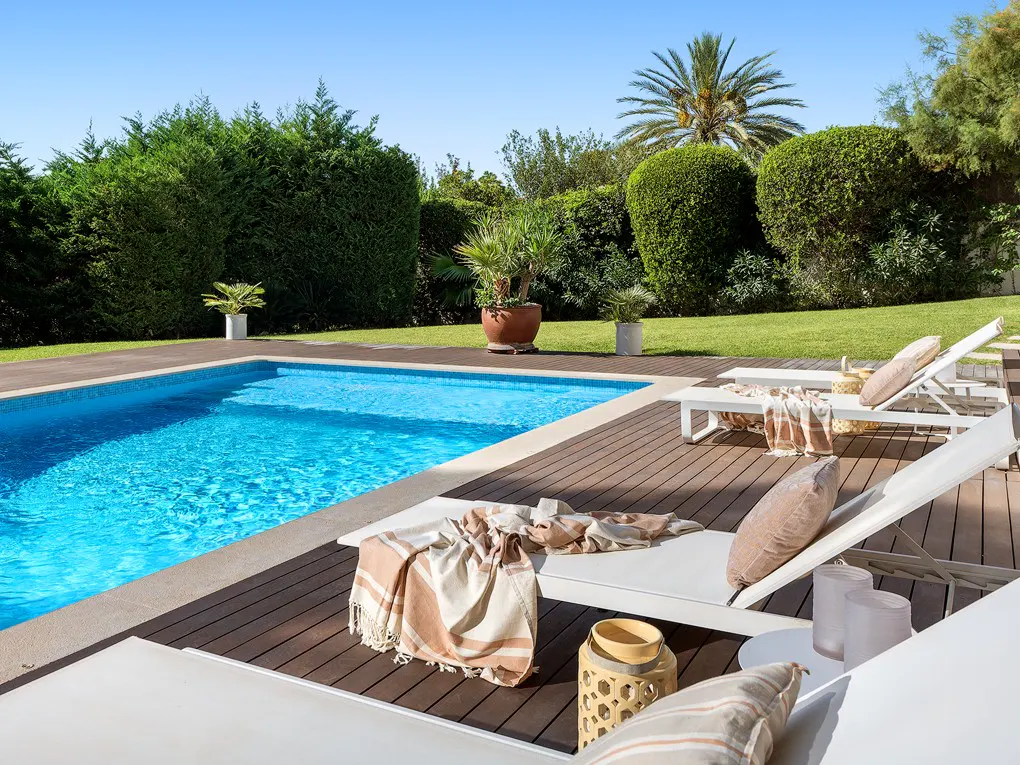 A backyard pool with lounge chairs on a wood deck, surrounded by green grass and trees.