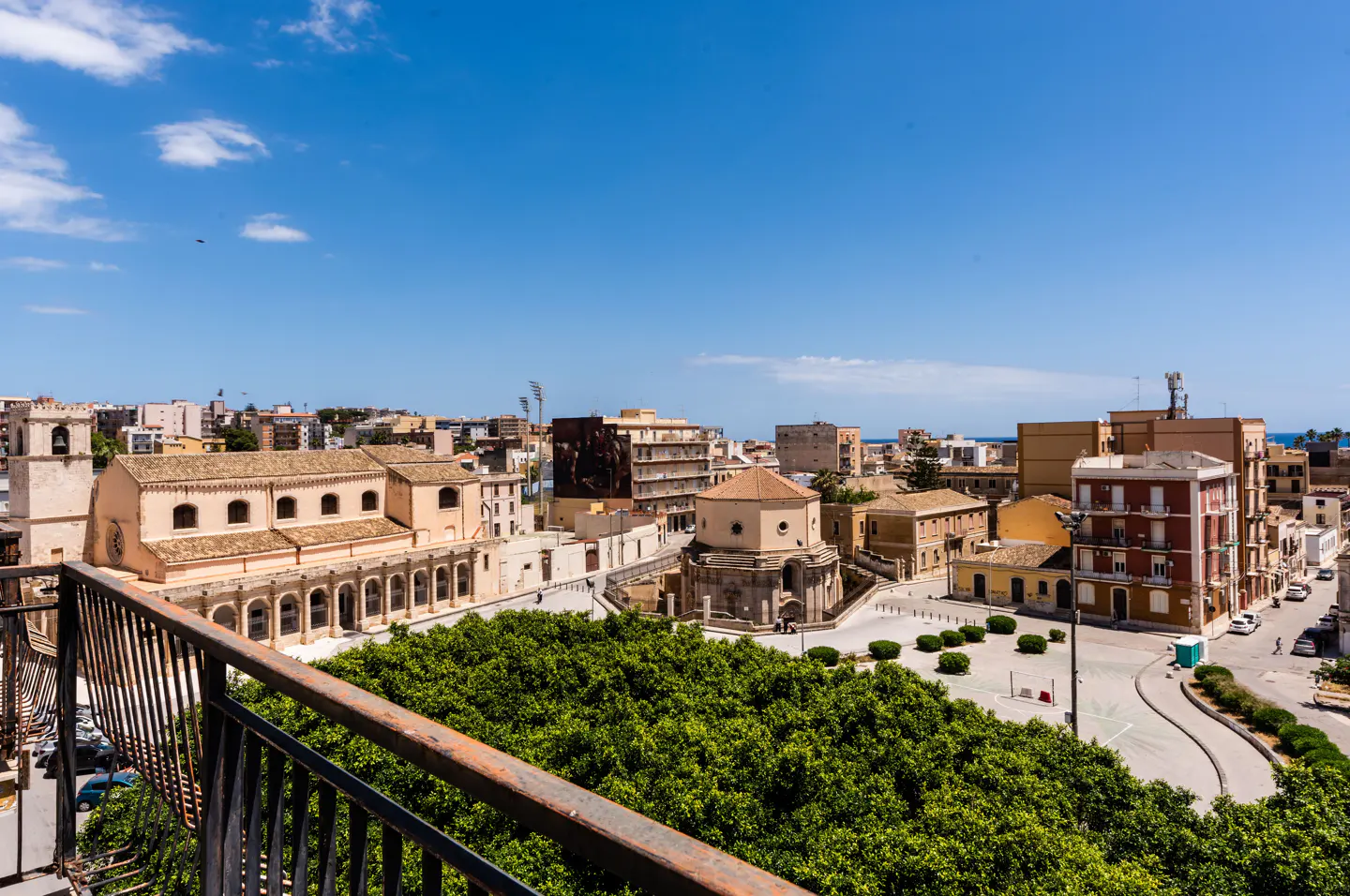 Elevated view of a sunlit Italian town square with historic stone buildings, a large green tree, and a clear blue sky.