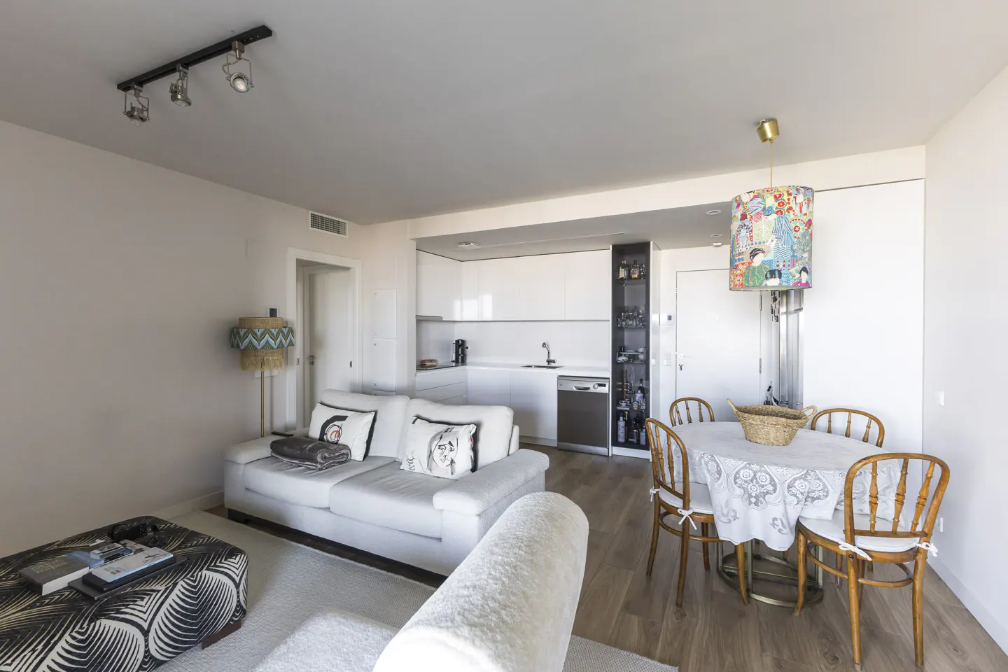 Bright, modern apartment interior with a white sofa, round dining table, and a compact kitchen in the background.
