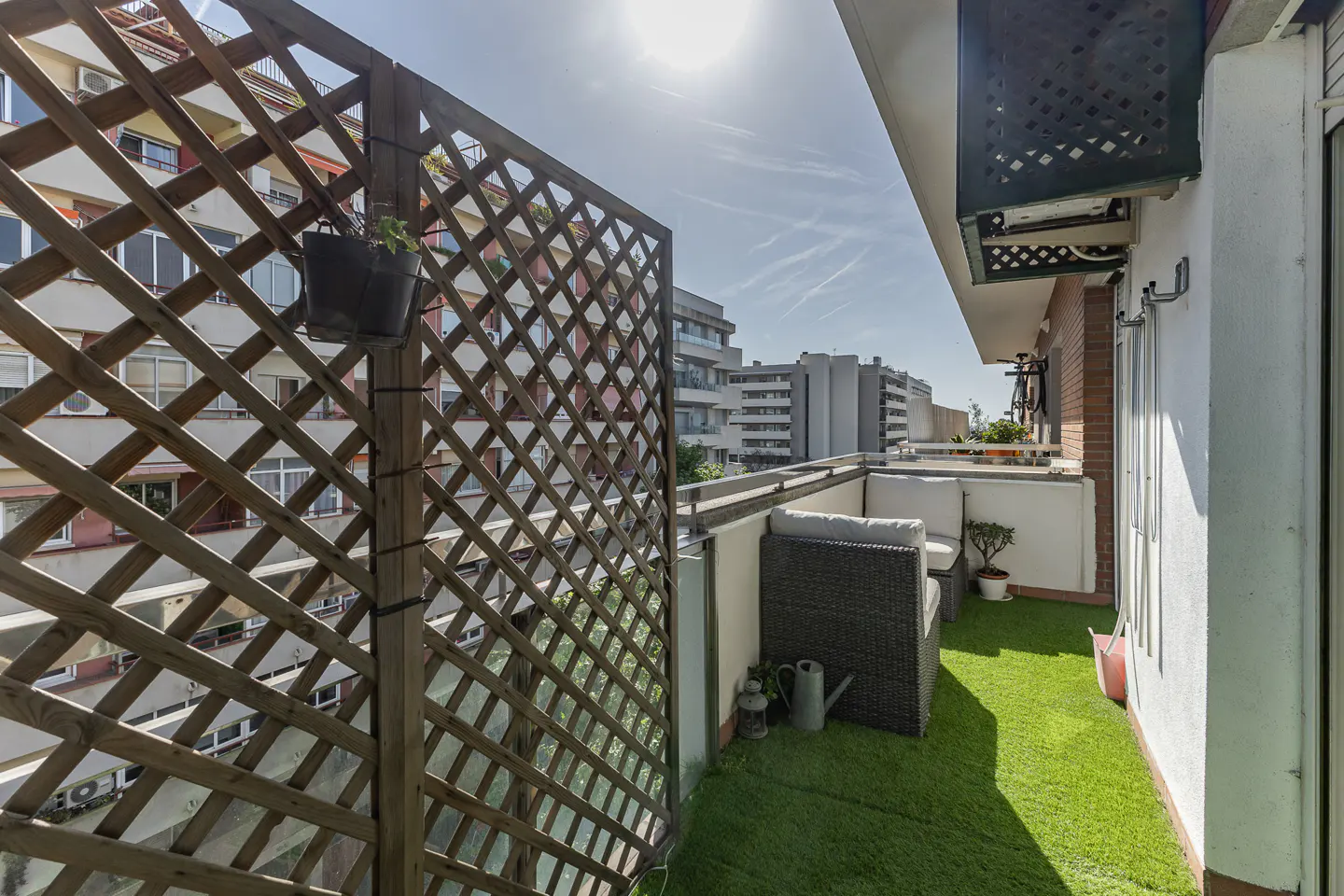 A sunny balcony with artificial grass, a wicker couch, and a wooden lattice screen. Buildings are visible in the background.