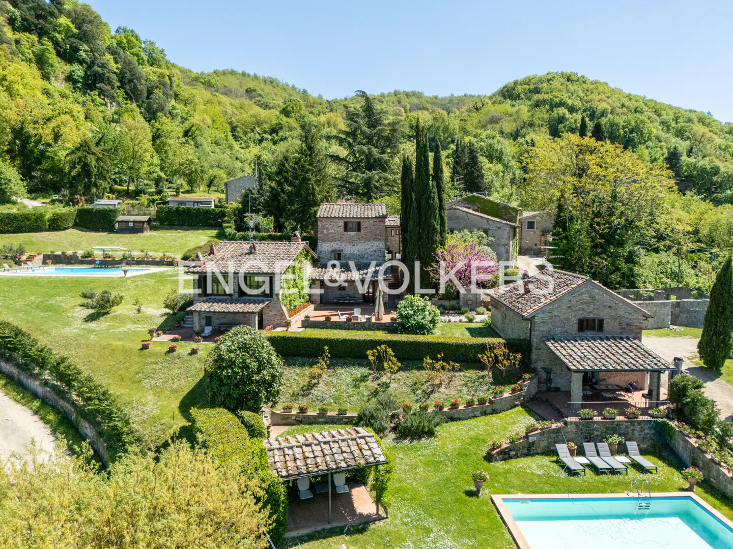 Aerial view of a stone house with a pool, surrounded by green hills and trees on a sunny day.