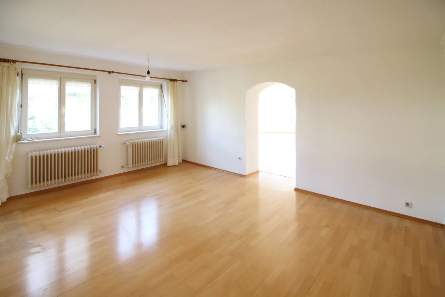 Empty room with light wood floors, white walls, two windows with white curtains, and an arched doorway.
