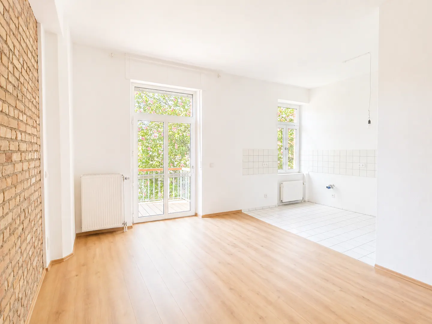 Bright, empty room with wood floors, white walls, and a brick accent wall. A balcony door and window let in natural light.