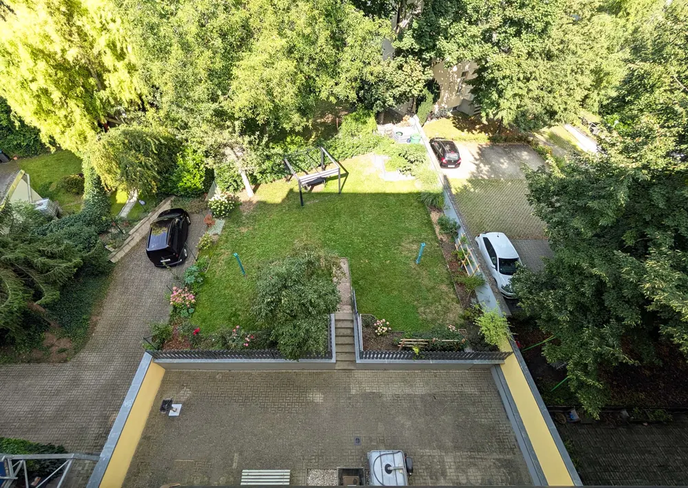 Aerial view of a backyard with green lawn, trees, patio, and parked cars. A swing set sits on the grass.