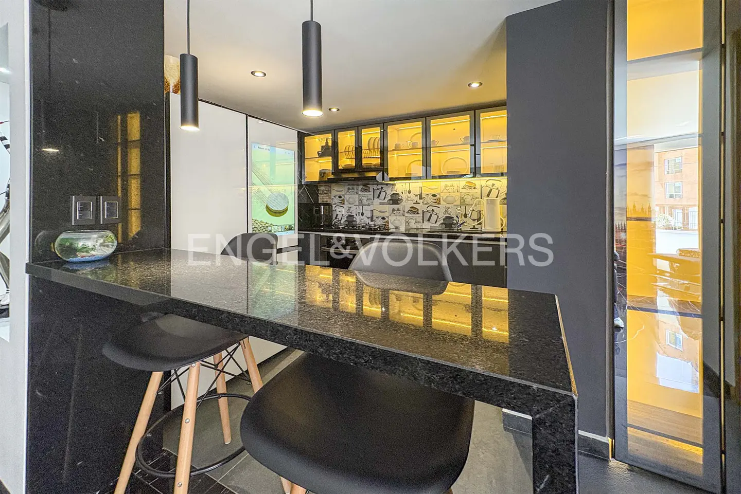 Modern kitchen with a black granite breakfast bar, two stools, and illuminated glass-front cabinets with a patterned backsplash.