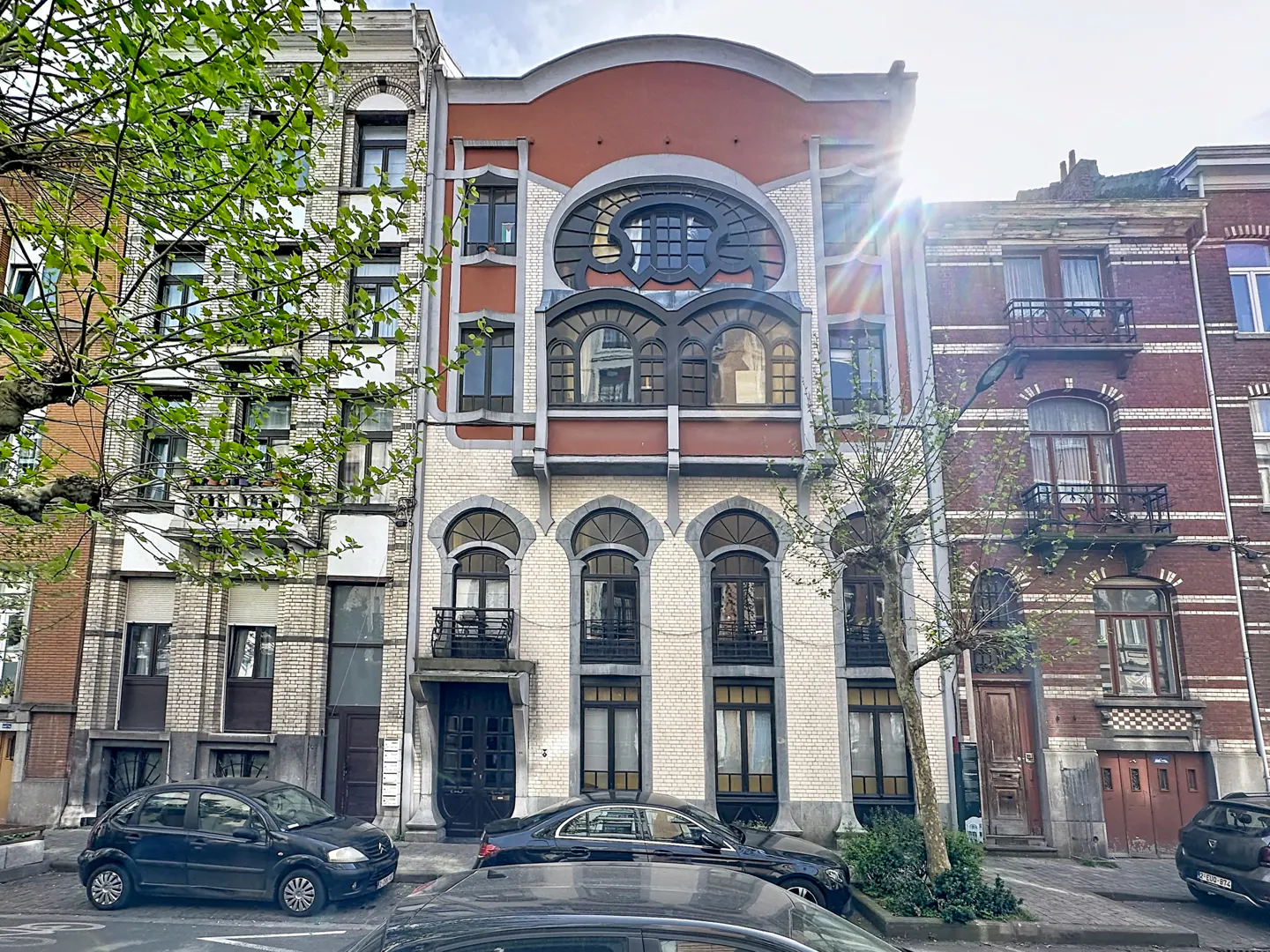Exterior view of a three-story building with white brick, arched windows, and a red trim. Cars are parked in front.