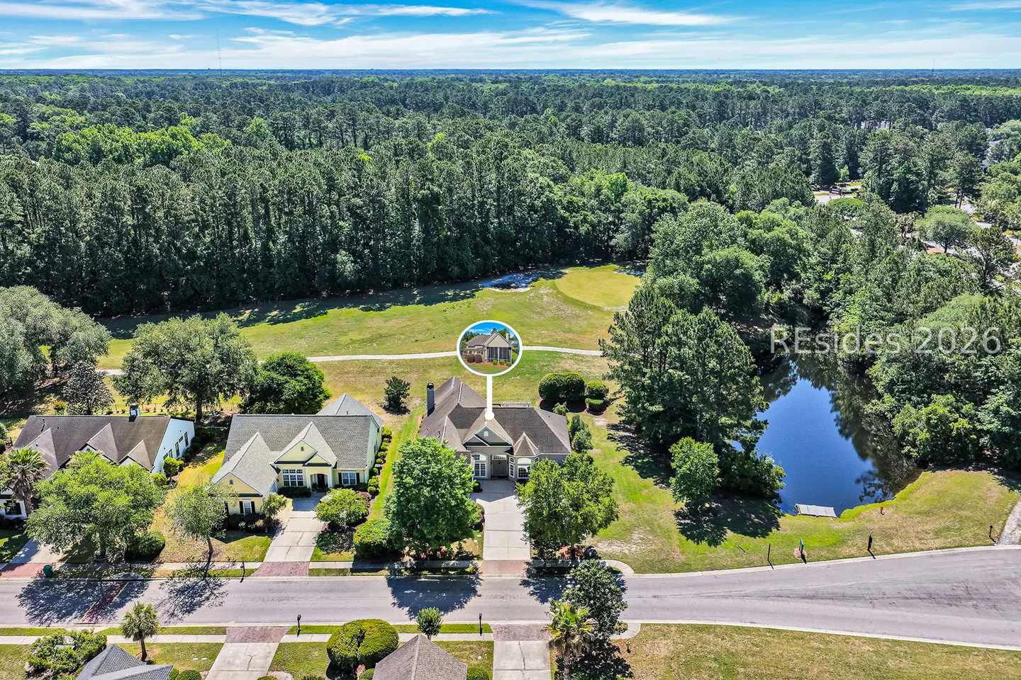 Aerial view of a tan house with a gray roof, green trees, a pond, and a golf course in the background.