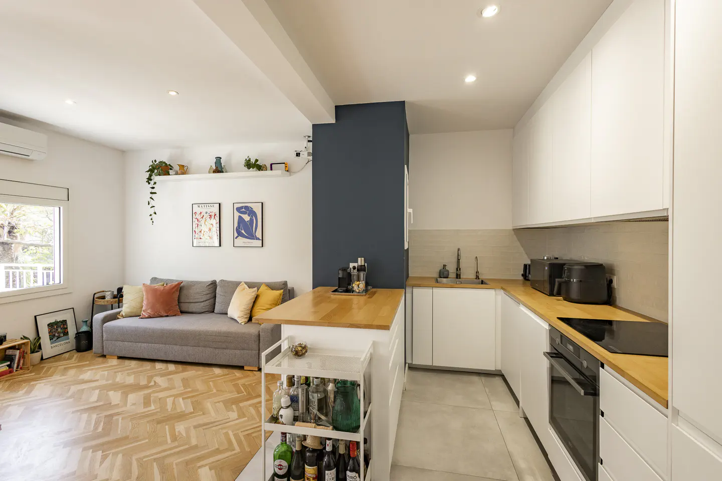 Open-concept apartment with a gray sofa, wood floors, and a white kitchen with wood countertops.