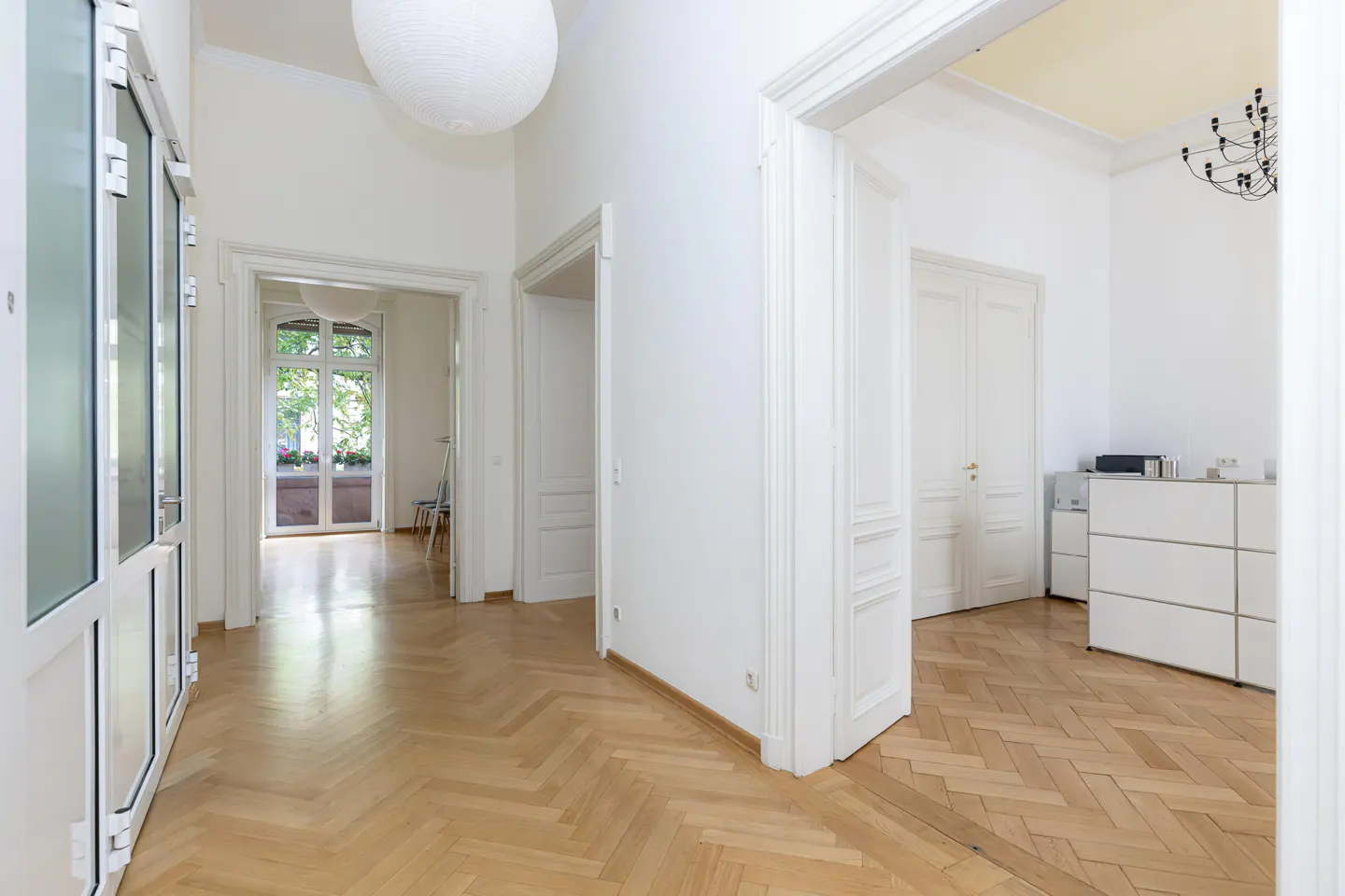 Bright hallway with herringbone wood floors, white walls, and multiple doorways. A large white paper lantern hangs from the ceiling.