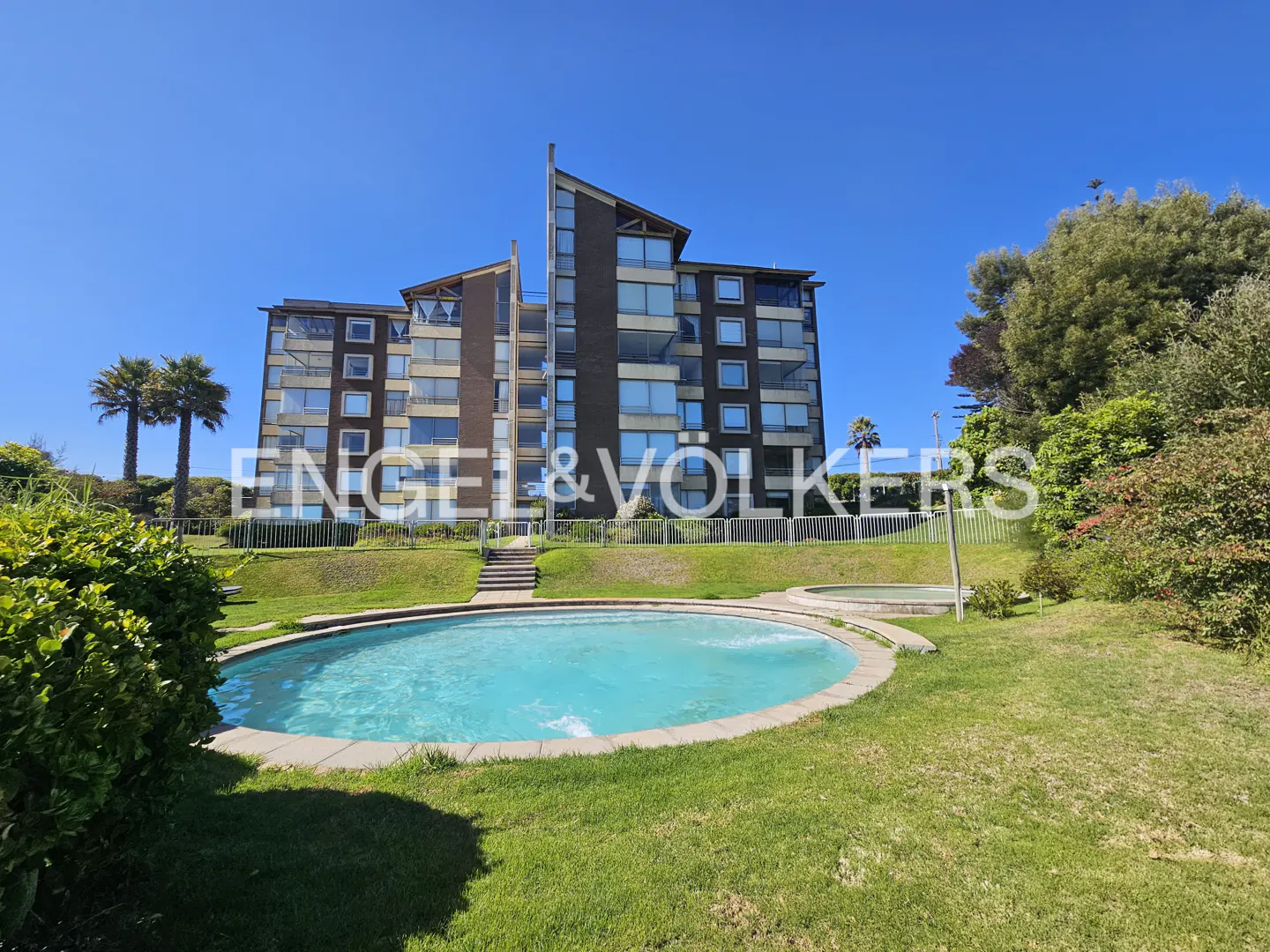Exterior view of a multi-story apartment building with a pool and green lawn under a clear blue sky. Palm trees are visible.