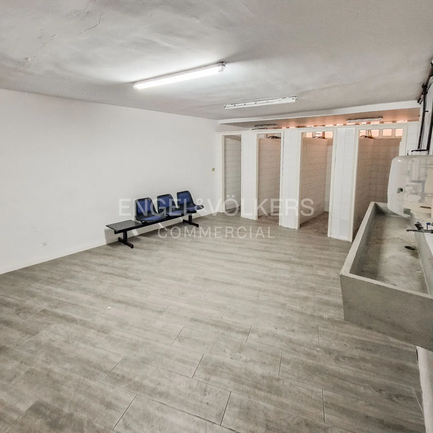 A bright, empty commercial space with blue seating, white walls, and a concrete sink. The floor is tiled in gray.