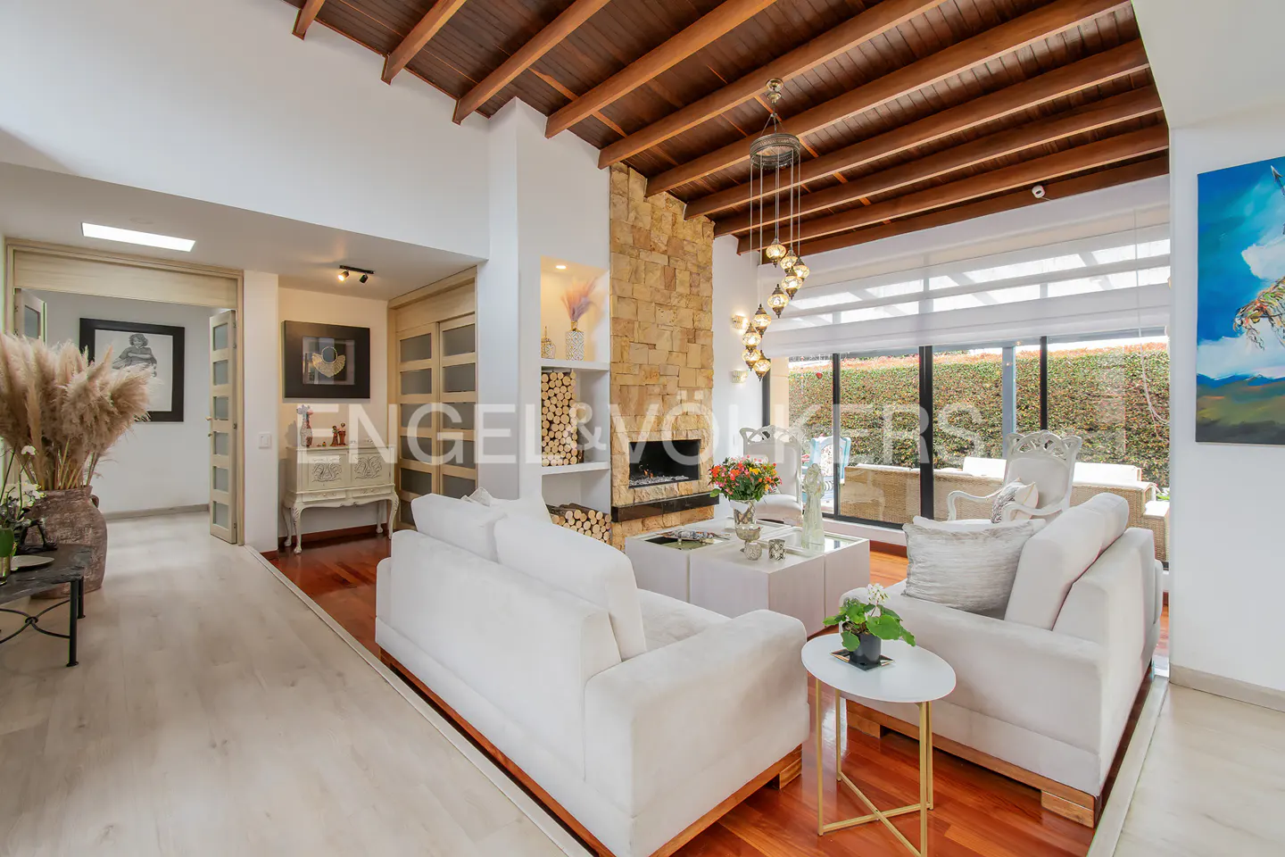 Bright living room with white sofas, wood floors, stone fireplace, and wood-beamed ceiling. Large windows overlook a green garden.