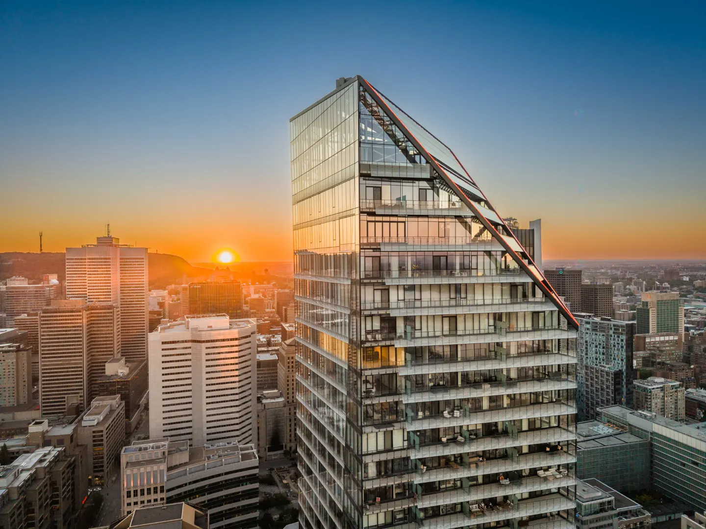 Modern glass skyscraper with a slanted roofline at sunset, overlooking a dense city skyline with warm golden light.