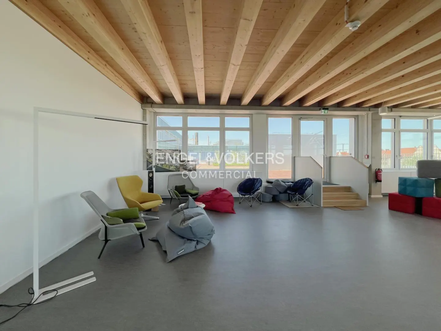 Bright, open office space with wooden beam ceiling, large windows, and colorful beanbag chairs. Engel & Völkers logo visible.