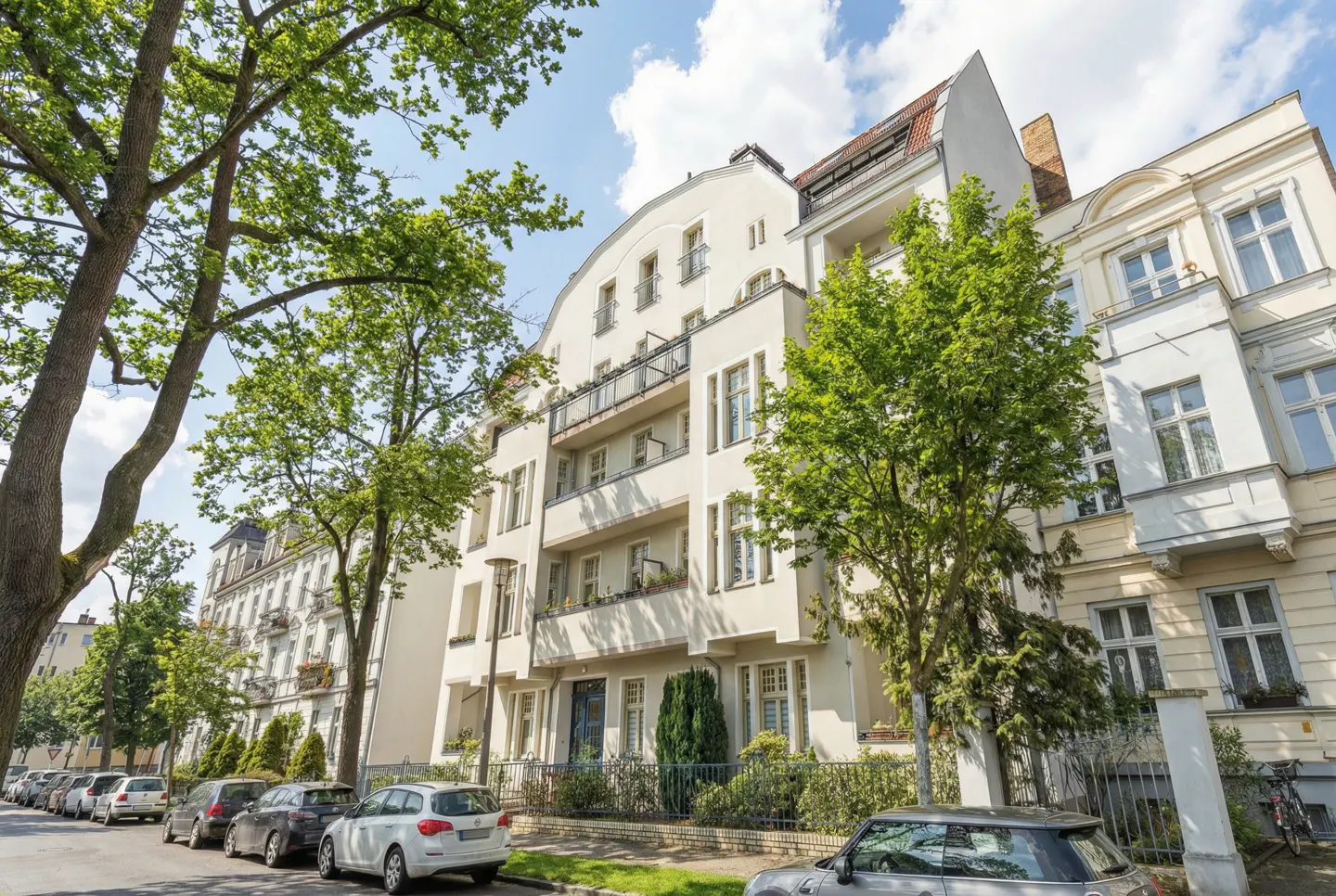 Street view of a cream-colored apartment building with balconies, green trees, and parked cars on a sunny day.