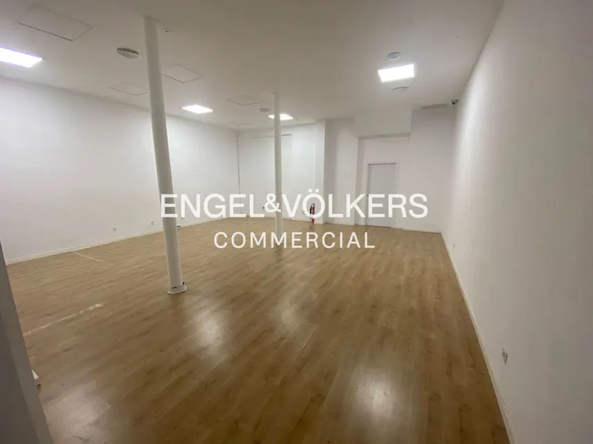 Bright, empty commercial space with light wood floors, white walls, and support columns. Engel & Völkers Commercial logo overlay.